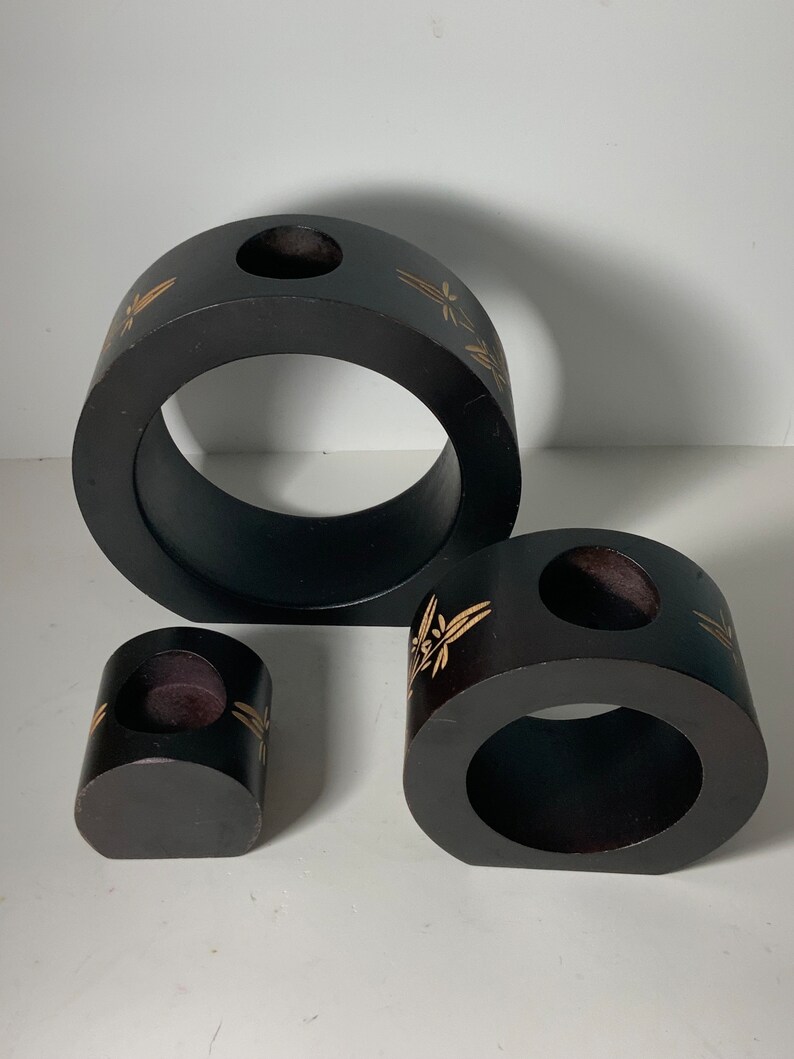 Puede incluir: Conjunto de tres candelabros de madera negra con dise&ntilde;os florales dorados. Los candelabros son de diferentes tama&ntilde;os y formas, con una abertura redonda en el centro de cada uno.