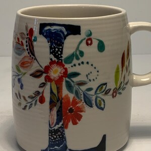 Puede incluir: Taza de cer&aacute;mica color crema con un dise&ntilde;o floral y la letra "L". La letra est&aacute; pintada en azul con un patr&oacute;n floral, complementado con flores y hojas rojas, naranjas y verdes. La taza tiene una forma curva y un asa.