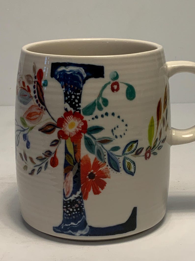 Puede incluir: Taza de cer&aacute;mica color crema con un dise&ntilde;o floral y la letra "L". La letra est&aacute; pintada en azul con un patr&oacute;n floral, complementado con flores y hojas rojas, naranjas y verdes. La taza tiene una forma curva y un asa.