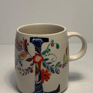 Puede incluir: Taza de cer&aacute;mica color crema con una gran "L" decorada con flores. La letra es principalmente azul marino, con acentos florales coloridos en rojo, naranja y verde. La taza tiene forma redondeada y un asa curva.