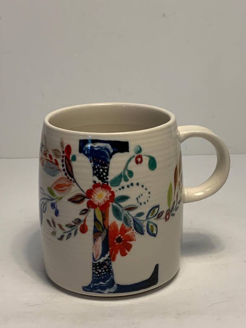 Puede incluir: Taza de cer&aacute;mica color crema con una gran "L" decorada con flores. La letra es principalmente azul marino, con acentos florales coloridos en rojo, naranja y verde. La taza tiene forma redondeada y un asa curva.
