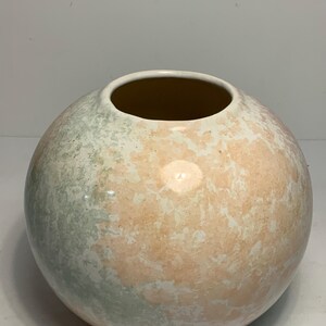 May include: A spherical ceramic vase with a wide opening. The vase features a speckled design with colors of peach, cream, and pale green. The interior of the vase is a warm brown color. The vase is a decorative item.