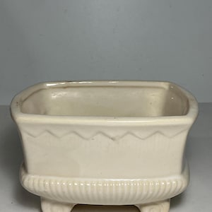 May include: A cream-coloured ceramic planter with a rectangular shape, featuring a horizontal zigzag pattern near the top rim and vertical ribbing along the base. The vessel is supported by four small, rounded feet at the bottom corners.