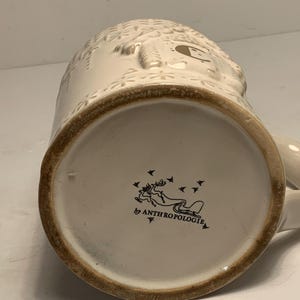Peut inclure: Mug en c&eacute;ramique cr&egrave;me avec des d&eacute;tails en relief et un bord marron. Le fond de la tasse pr&eacute;sente un logo avec un renne tirant un tra&icirc;neau et le texte "Anthropologie". La tasse a une anse.
