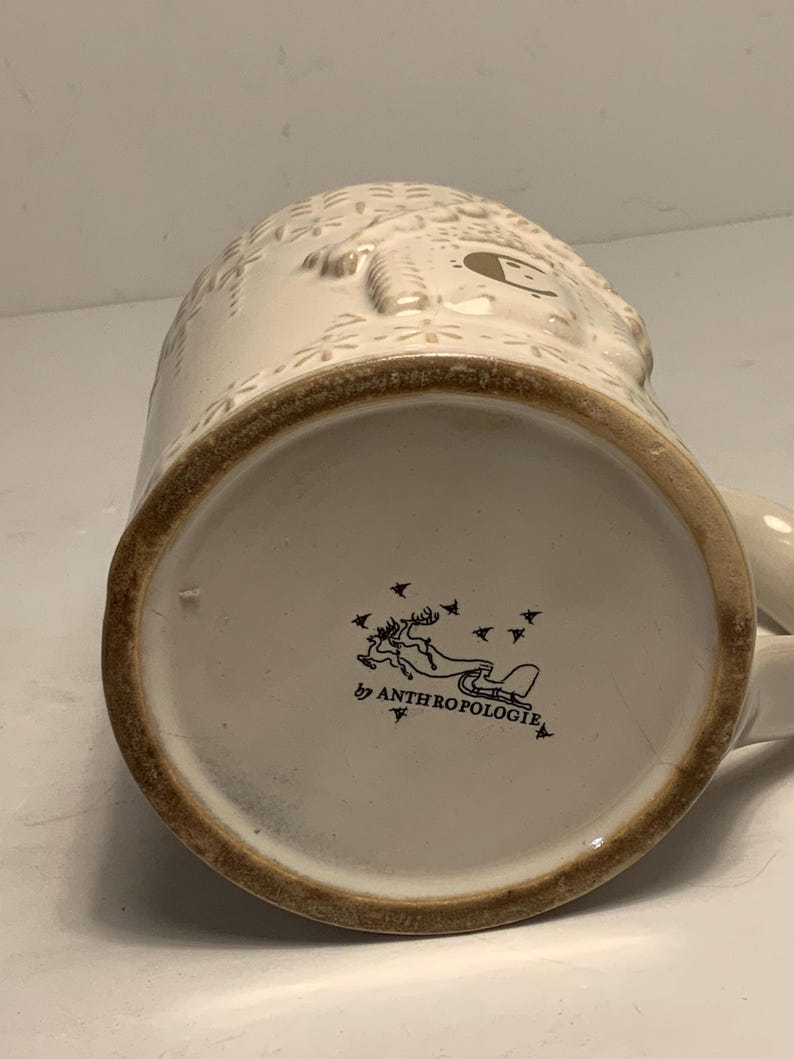 Peut inclure: Mug en c&eacute;ramique cr&egrave;me avec des d&eacute;tails en relief et un bord marron. Le fond de la tasse pr&eacute;sente un logo avec un renne tirant un tra&icirc;neau et le texte "Anthropologie". La tasse a une anse.