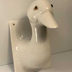 May include: A white ceramic duck head wall hanging with a rectangular base. The duck has yellow eyes and a white beak. The duck's neck and body are detailed with textured feathers. The piece is mounted on a white rectangular base with two holes.