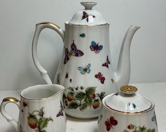 Op de afbeelding: Een wit porseleinen theeservies bestaande uit een theepot, een melkkannetje en een suikerpot met deksel. Elk stuk heeft een geribbelde textuur, goudkleurige randen en een kleurrijk patroon van diverse vlinders en rode aardbeien.
