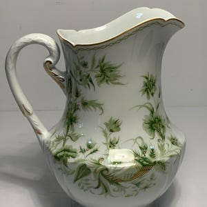 Könnte beinhalten: Weißer Porzellan-Krug mit gebogenem Henkel und ausgestelltem Ausguss. Der Krug ist mit grünen Blumenmustern und goldenen Akzenten verziert. Der Rand hat eine zarte Goldverzierung. Der Krug hat einen bauchigen Boden.