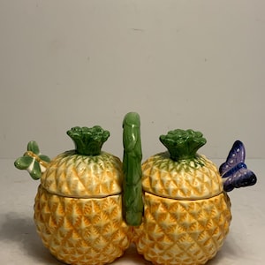 Könnte beinhalten: Ein Keramik-Gewürzset in Ananasform mit grünem Griff und Deckeln. Die gelb-cremefarbenen Ananastöpfe haben grüne Oberteile und sind mit Schmetterlingsakzenten verziert. Das Set ist für Salz und Pfeffer oder andere Gewürze konzipiert.