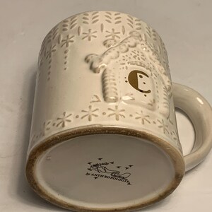 Peut inclure: Mug en c&eacute;ramique cr&egrave;me avec un motif en relief de maison en pain d'&eacute;pices et des flocons de neige. La tasse a un bord et une anse marron. Le fond de la tasse porte la mention "Anthropologie".