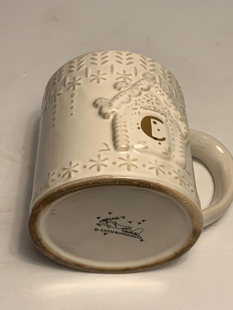 Peut inclure: Mug en c&eacute;ramique cr&egrave;me avec un motif en relief de maison en pain d'&eacute;pices et des flocons de neige. La tasse a un bord et une anse marron. Le fond de la tasse porte la mention "Anthropologie".