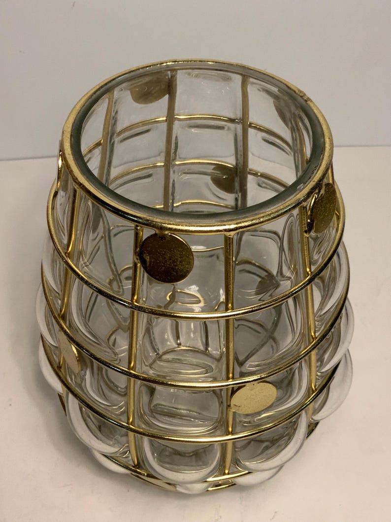 Op de afbeelding: Een heldere glazen vaas met een gouden metalen kooi-ontwerp. De vaas heeft een afgeronde vorm met een textuur, bubbelachtig patroon. De gouden kooi is gemaakt van verticale en horizontale staven, met kleine gouden schijfaccenten.