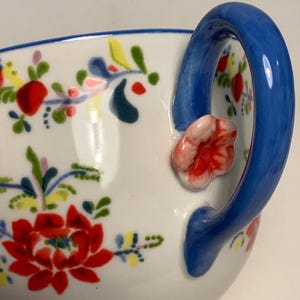 May include: White ceramic teacup with a blue handle and floral design. The cup features a vibrant red flower with green leaves and accents of yellow and blue. A small pink flower is attached to the handle.