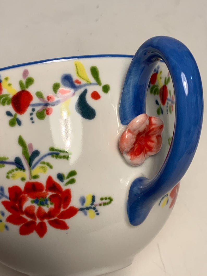 May include: White ceramic teacup with a blue handle and floral design. The cup features a vibrant red flower with green leaves and accents of yellow and blue. A small pink flower is attached to the handle.