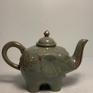 Puede incluir: Una tetera de cerámica con forma de elefante, con un esmalte gris verdoso y detalles marrones. La tetera tiene un asa curvada, un pico con forma de trompa de elefante y una tapa con un pequeño pomo. El cuerpo del elefante está decorado con patrones en relieve.