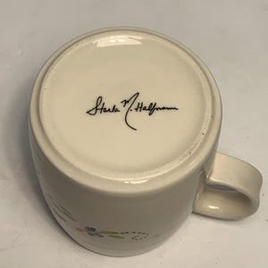 Puede incluir: Taza de cer&aacute;mica blanquecina con asa curvada. La base de la taza presenta la firma "Starla M. Halfmann" en escritura negra. La taza tiene un dise&ntilde;o sencillo con peque&ntilde;os detalles florales en tonos azules, verdes y amarillos.