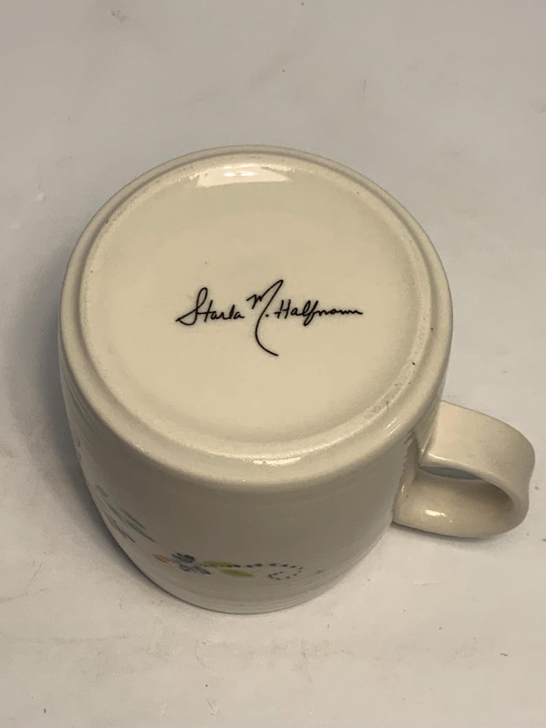 Puede incluir: Taza de cer&aacute;mica blanquecina con asa curvada. La base de la taza presenta la firma "Starla M. Halfmann" en escritura negra. La taza tiene un dise&ntilde;o sencillo con peque&ntilde;os detalles florales en tonos azules, verdes y amarillos.