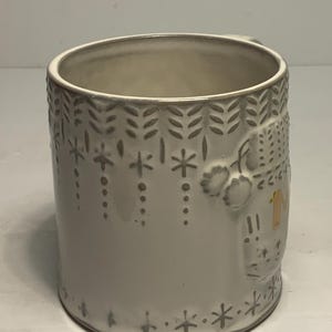 May include: A white ceramic mug with embossed winter-themed designs. The mug features a handle and is decorated with snowflakes, leaves, and a house. The interior of the mug is a light beige color.