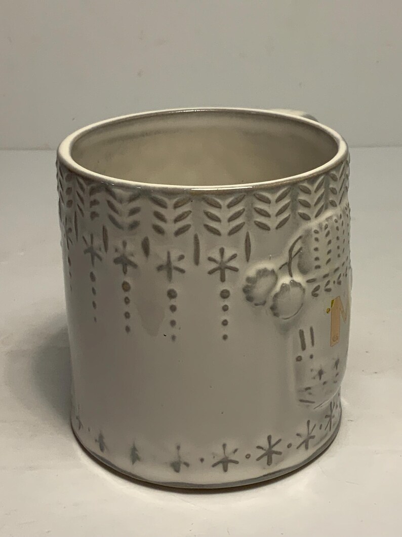 May include: A white ceramic mug with embossed winter-themed designs. The mug features a handle and is decorated with snowflakes, leaves, and a house. The interior of the mug is a light beige color.