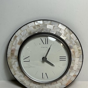 May include: A round wall clock featuring a frame composed of small, iridescent white mother-of-pearl tiles. The clock face is off-white with black Roman numerals at the twelve, three, six, and nine o'clock positions, the word Domicile, and two hands.