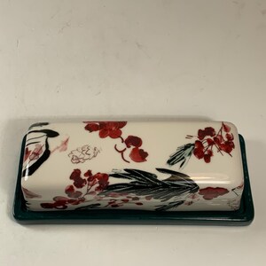 May include: A rectangular butter dish with a white lid featuring a floral design in red and black. The base is a dark green. The dish is designed to hold a standard size of butter.