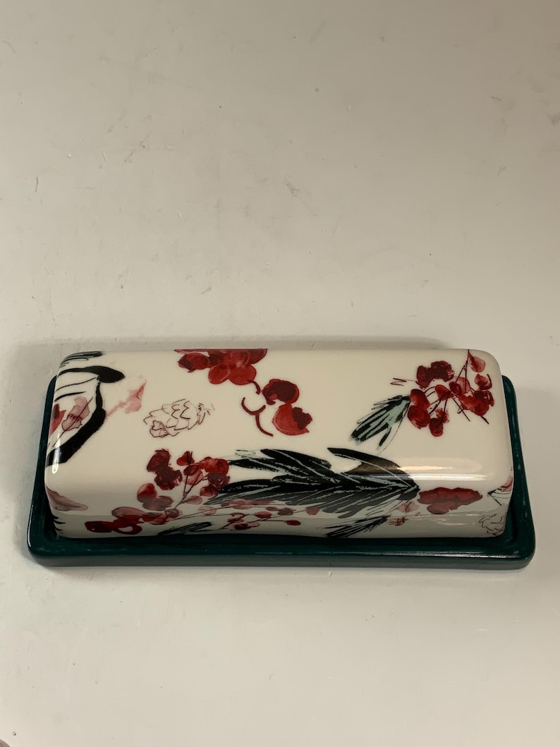 May include: A rectangular butter dish with a white lid featuring a floral design in red and black. The base is a dark green. The dish is designed to hold a standard size of butter.