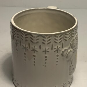 May include: A white ceramic mug with a handle. The mug features a repeating pattern of snowflakes, dots, and leaf-like shapes in a light gray color. The mug has a slightly textured surface.