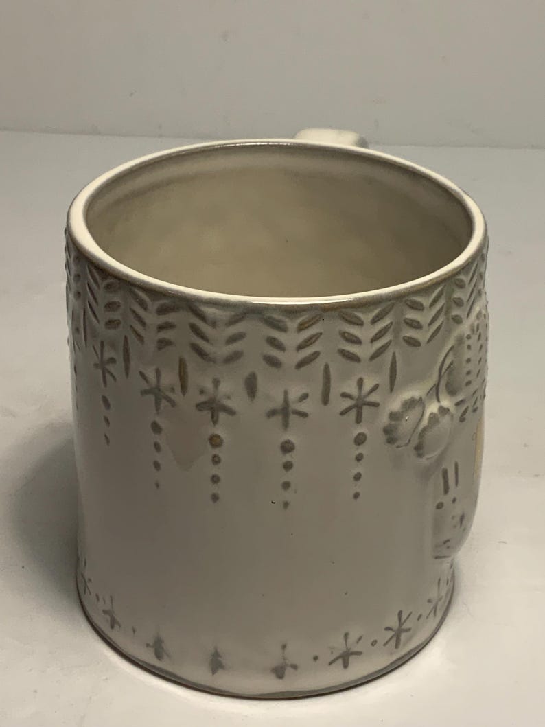 May include: A white ceramic mug with a handle. The mug features a repeating pattern of snowflakes, dots, and leaf-like shapes in a light gray color. The mug has a slightly textured surface.