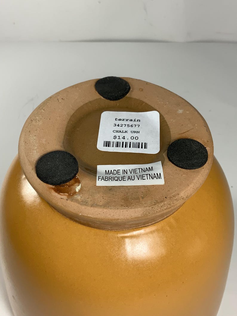 Pu&ograve; includere: Un'urna in ceramica marrone con un'etichetta che recita "terrain 34275677 CHALK URN $ 14.00" e "MADE IN VIETNAM FABRIQUE AU VIETNAM".