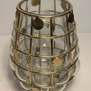 Op de afbeelding: Transparante glazen vaas met een gouden metalen kooi-ontwerp. De vaas heeft een afgeronde vorm met een reeks belachtige glazen secties. Goudkleurige cirkelvormige accenten zijn bevestigd aan de metalen kooi. De vaas is decoratief.