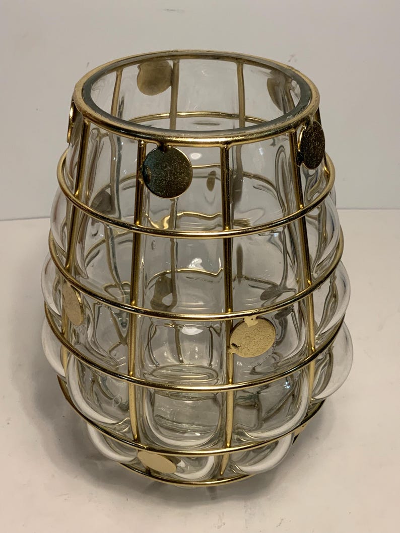 Op de afbeelding: Transparante glazen vaas met een gouden metalen kooi-ontwerp. De vaas heeft een afgeronde vorm met een reeks belachtige glazen secties. Goudkleurige cirkelvormige accenten zijn bevestigd aan de metalen kooi. De vaas is decoratief.