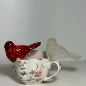 May include: A white teacup with a floral pattern, adorned with a red glass bird and a frosted glass bird. The teacup has a gold rim and a handle. The floral design includes pink roses and green leaves.