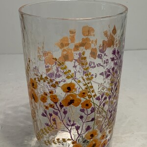 May include: A clear glass tumbler with a textured surface. The glass is decorated with a floral design in shades of orange and purple. The flowers and foliage are painted on the glass, creating a decorative drinking glass.