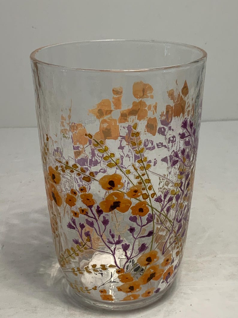 May include: A clear glass tumbler with a textured surface. The glass is decorated with a floral design in shades of orange and purple. The flowers and foliage are painted on the glass, creating a decorative drinking glass.