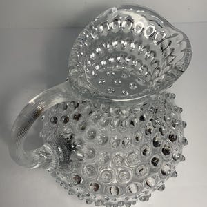 May include: Clear glass pitcher with a textured, hobnail design. The pitcher features a handle and a spout for pouring. The hobnail pattern covers the body of the pitcher, creating a decorative and tactile surface.