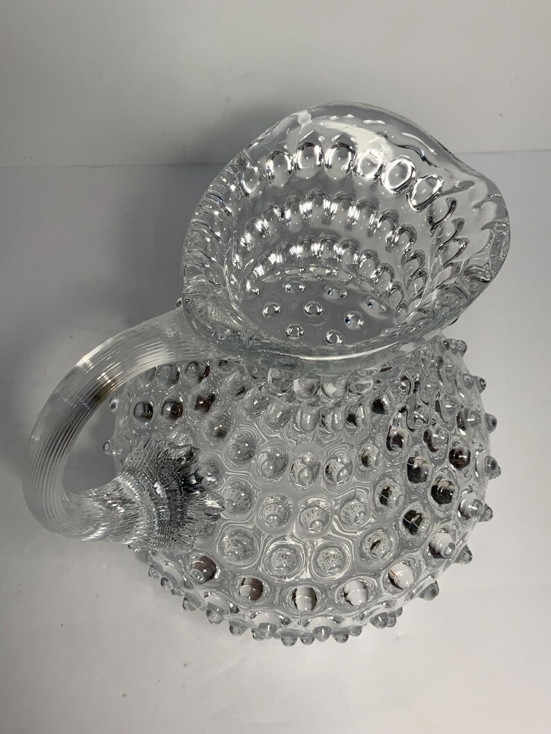 May include: Clear glass pitcher with a textured, hobnail design. The pitcher features a handle and a spout for pouring. The hobnail pattern covers the body of the pitcher, creating a decorative and tactile surface.