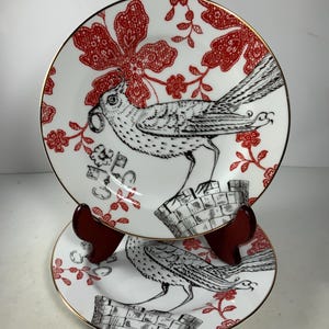 May include: Two white ceramic plates with a red and black floral design. The plates feature a black and white bird with a long tail. The plates have a gold rim.