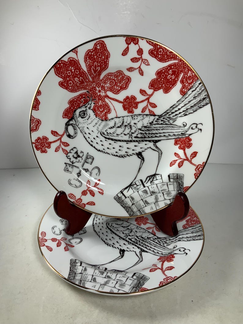 May include: Two white ceramic plates with a red and black floral design. The plates feature a black and white bird with a long tail. The plates have a gold rim.