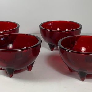May include: Four ruby red glass bowls with a rounded shape and three small legs. The bowls have a glossy finish and are arranged on a white surface. These bowls are suitable for serving snacks or small portions.