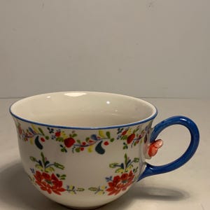 May include: White ceramic teacup with a blue handle and rim. The cup is decorated with red and yellow floral patterns. A small, red floral accent is on the handle. The cup is designed for tea or coffee.