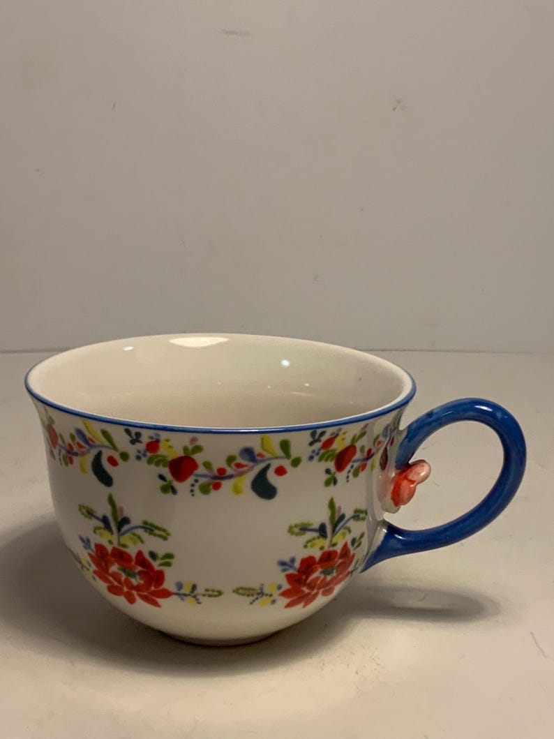 May include: White ceramic teacup with a blue handle and rim. The cup is decorated with red and yellow floral patterns. A small, red floral accent is on the handle. The cup is designed for tea or coffee.
