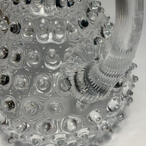May include: A clear glass pitcher with a textured, bubble-like surface. The pitcher has a large, curved handle and a wide base. The glass is transparent, allowing light to pass through. The design is decorative and unique.