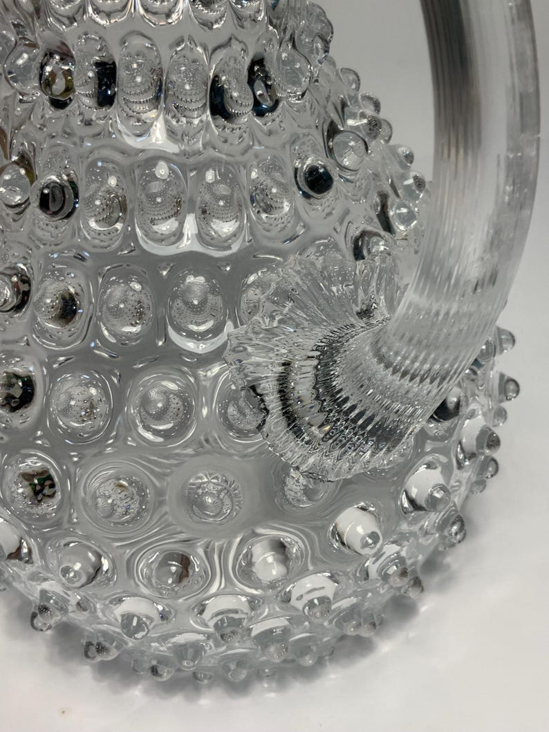 May include: A clear glass pitcher with a textured, bubble-like surface. The pitcher has a large, curved handle and a wide base. The glass is transparent, allowing light to pass through. The design is decorative and unique.
