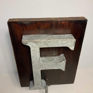 May include: A metal letter "F" mounted on a dark wood plaque, supported by a metal stand. The letter has a weathered, industrial appearance. The wood has a rich, dark brown color. The stand is square and made of the same metal as the letter.