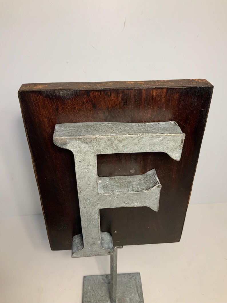 May include: A metal letter "F" mounted on a dark wood plaque, supported by a metal stand. The letter has a weathered, industrial appearance. The wood has a rich, dark brown color. The stand is square and made of the same metal as the letter.