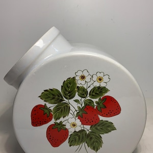 Pode incluir: Recipiente de cerâmica branca com um design curvo e uma abertura inclinada. O recipiente é decorado com um motivo de morango, com morangos vermelhos, folhas verdes e flores brancas.
