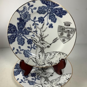 May include: Two white porcelain plates with a blue and black floral design and a deer illustration. The plates have a gold rim and a coat of arms with a crown and a lion.