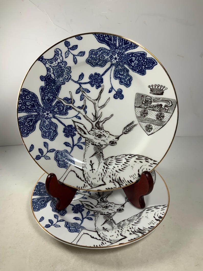 May include: Two white porcelain plates with a blue and black floral design and a deer illustration. The plates have a gold rim and a coat of arms with a crown and a lion.