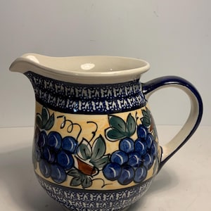 Puede incluir: Jarra de cerámica con borde y asa color crema, y diseño azul y blanco. La jarra presenta un diseño pintado a mano de uvas azules y hojas verdes sobre fondo amarillo. La jarra mide aproximadamente 20 cm de altura.
