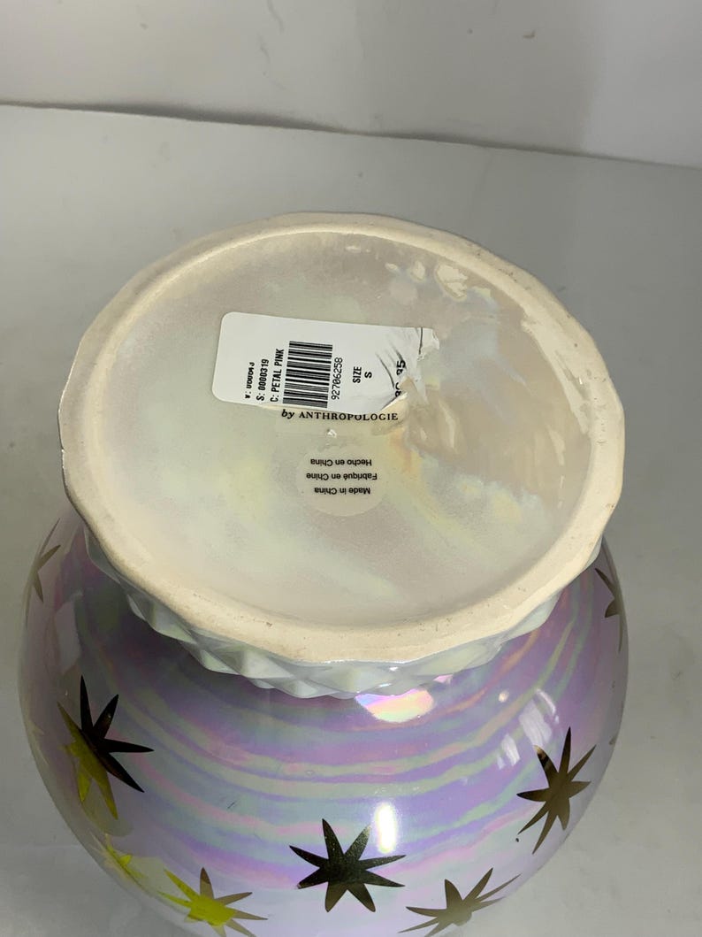 May include: A pearlescent vase with a light purple and gold star design. The vase has a wide opening and a slightly ruffled rim. The label reads "by Anthropologie".